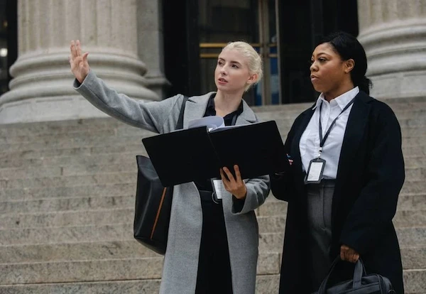 Two women in professional suits stand on steps, holding a folder