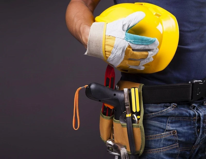 . A construction worker with a hard hat and safety glasses holds a tool belt